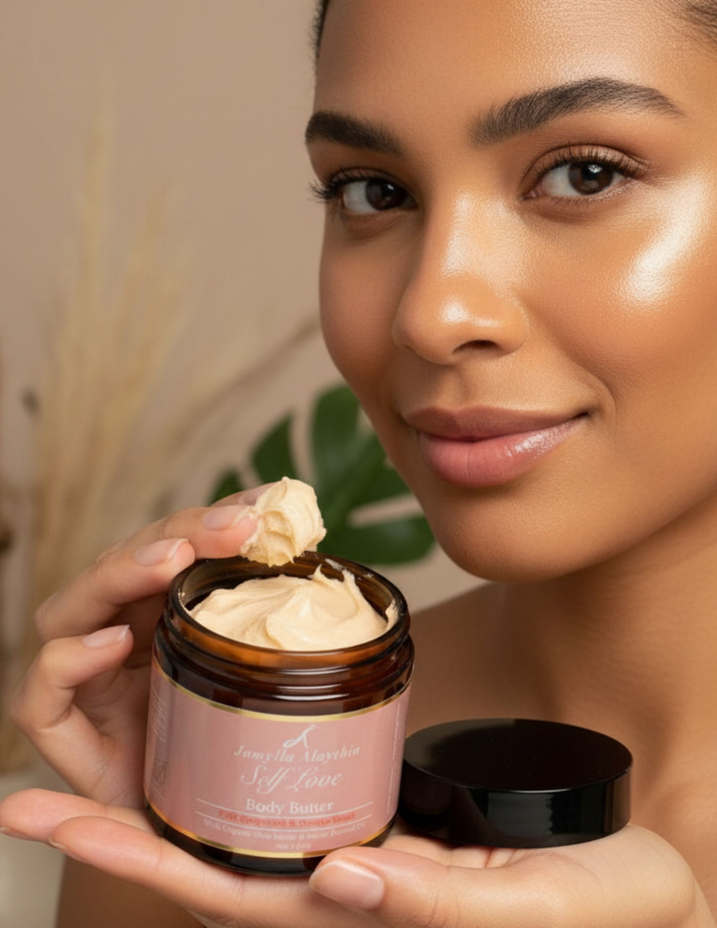 Woman holding a jar of self love body butter with the lid off and the whipped butter showing. she has some of the body butter on her fingers.A visible brand name, which says 'Jamylla Alaythia'