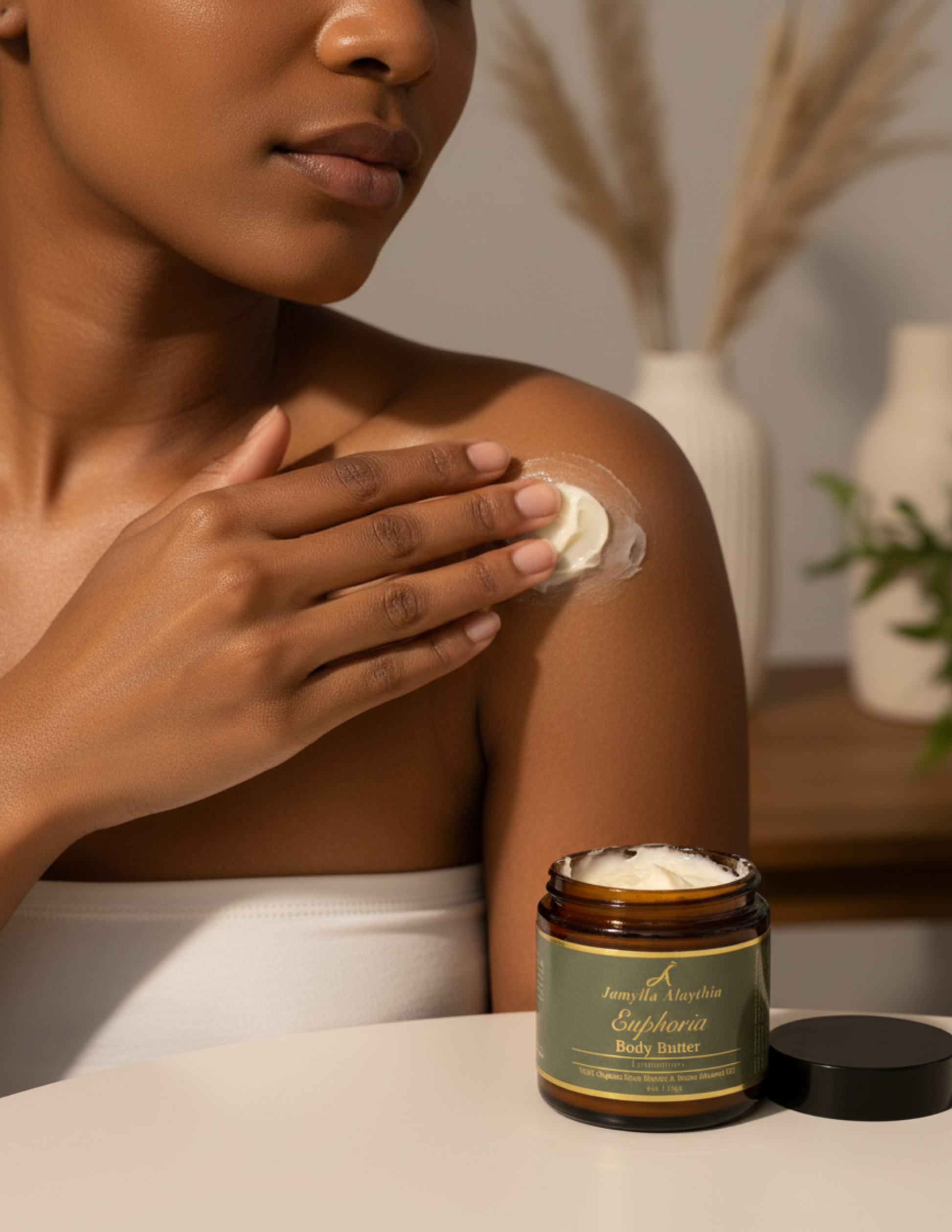 Woman applying Euphoria Whipped body butter to her shoulder with the amber glass jar and a green and gold label with the black lid off on a table in the foreground.  