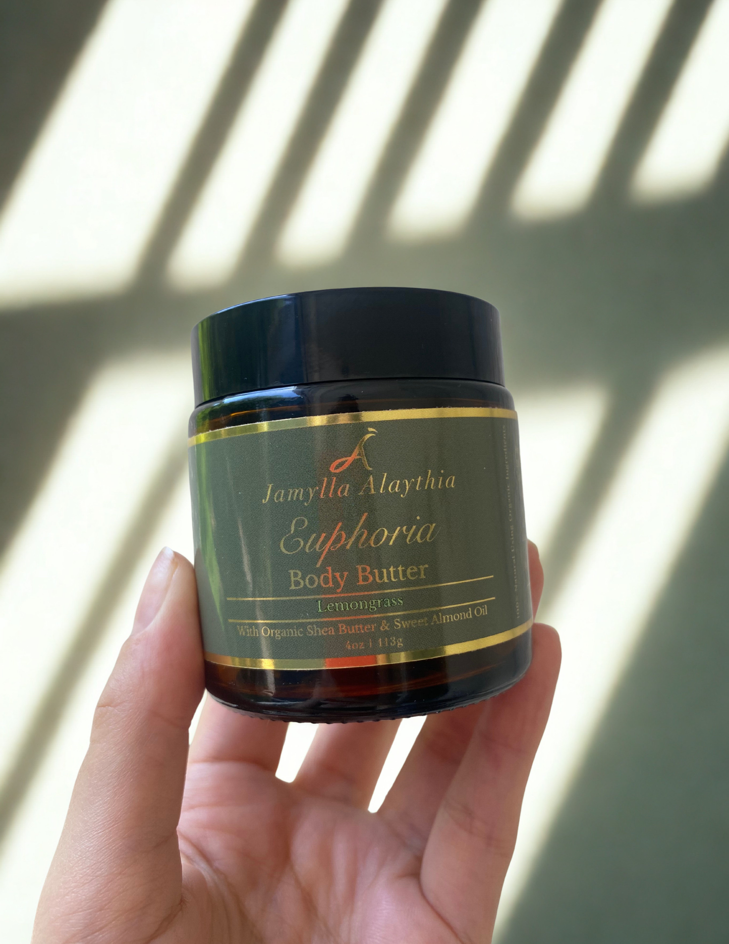 Hand holding amber glass jar green lid of Jamylla Alaythia Euphoria Body Butter with a sunlit green background