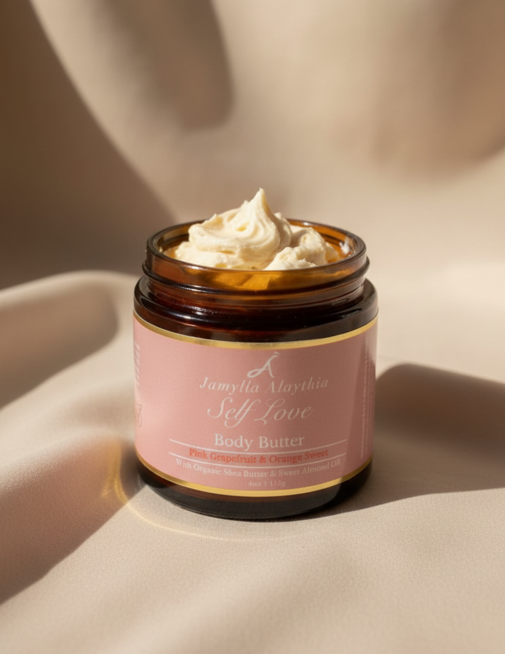 Jar of self love body butter, lid off, creamy whipped butter visible, with a pink label on a beige background of a soft material