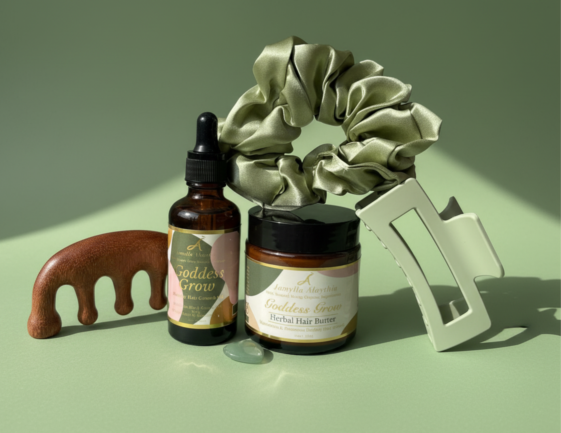 Set of hair care products including an amber glass bottle os Goddess Grow Hair Growth Oil , an amber glass jar of Goddess Grow Herbal Hair Butter, a woden sandalwood comb, a sage green satin crunchie and sage green claw clip a  on a sage green background with soft shadows